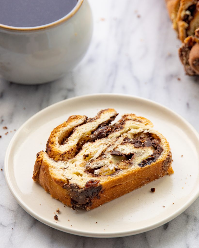 a slice of babka on a plate on a marble surface with a cup of coffee in the background