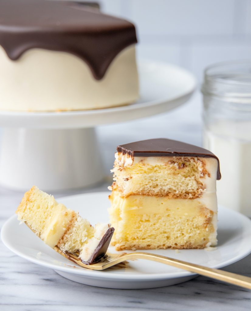 a slice of Boston Cream Pie on a plate with one bite on a fork and the rest of the cake in the background