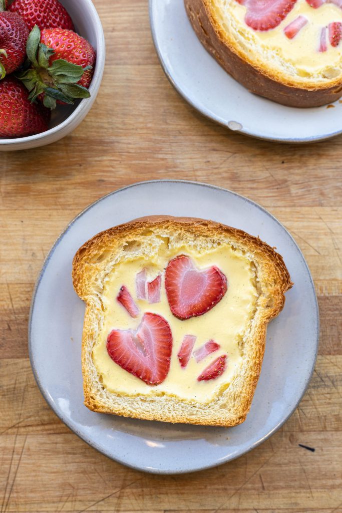 A piece of custard toast with sliced strawberries on top sits on a plate next to another plate and a bowl of strawberries.