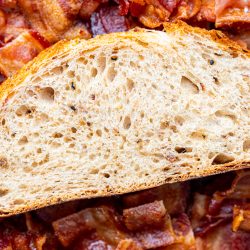 A slice of peppered bacon farm bread lays on a bed of chopped bacon.