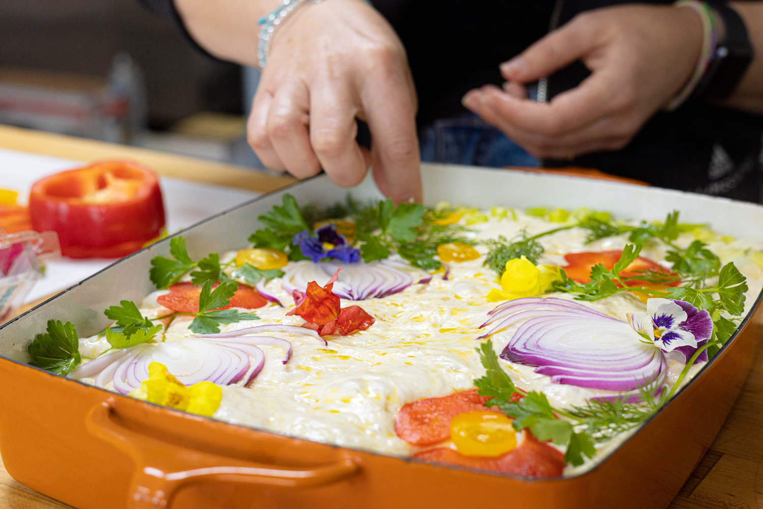 A pair of hands are seen adding toppings to the unbaked focaccia dough