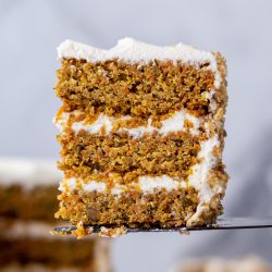 A slice of carrot cake is being lifted on a cake server with the remainder of the cake out of focus in the background.