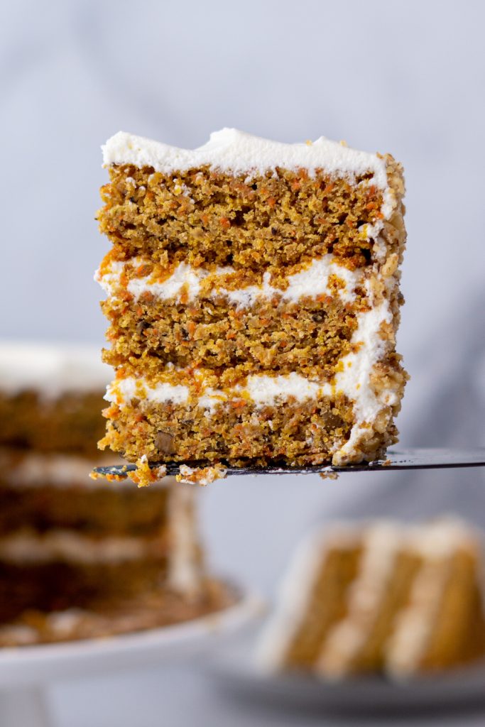 A slice of carrot cake is being lifted on a cake server with the remainder of the cake out of focus in the background.