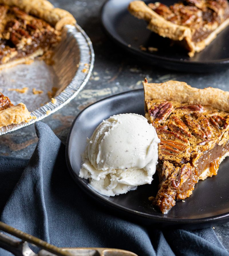 a slice of pecan pie on a black plate with a scoop of vanilla ice cream with the rest of the pie in the background
