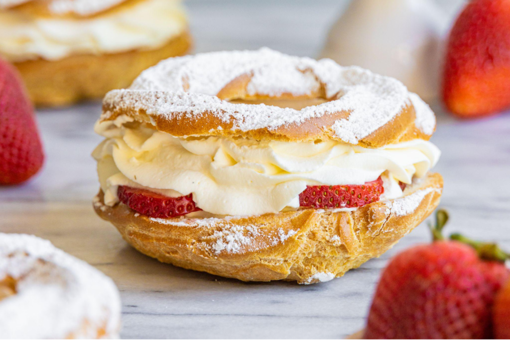 A strawberry Paris Brest sits on a marble surface with more Paris Brest and whole strawberries around it