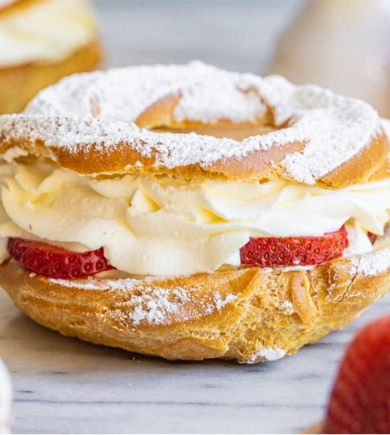 A strawberry Paris Brest sits on a marble surface with more Paris Brest and whole strawberries around it
