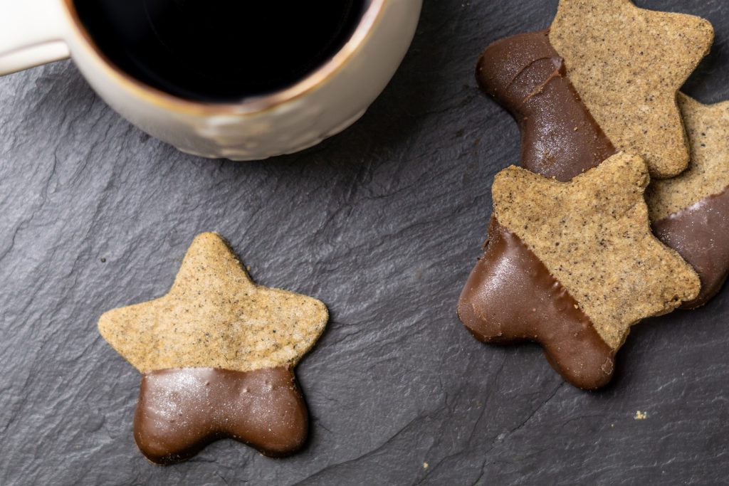 4 Chocolate-Dipped Espresso Star cookies strewn about on a black slate surface with a cream ceramic mug of coffee visible but out of focus at the top.
