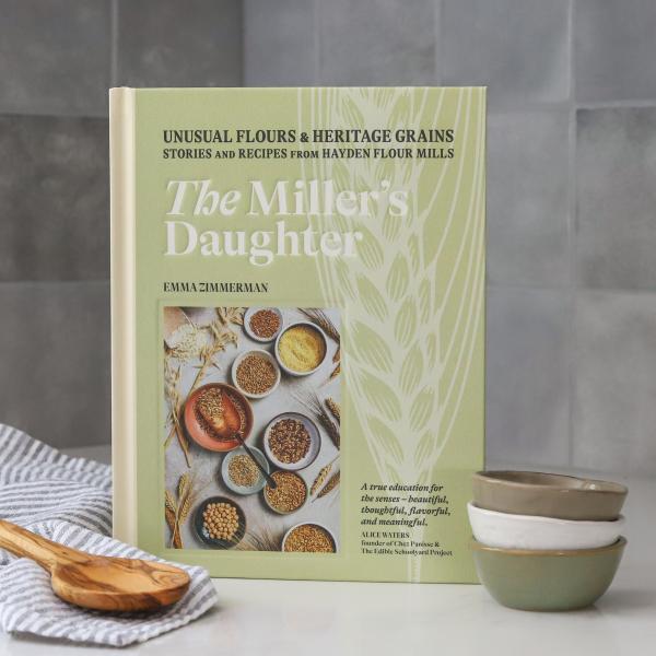 The Miller's Daughter cookbook standing upright with 3 small bowl and a wooden spoon alongside it