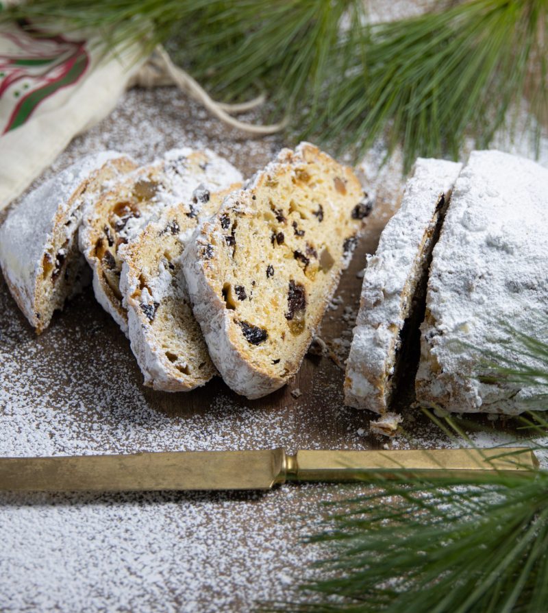 a loaf of stollen on a powdered sugar-dusted surface, with a gold knife and greenery