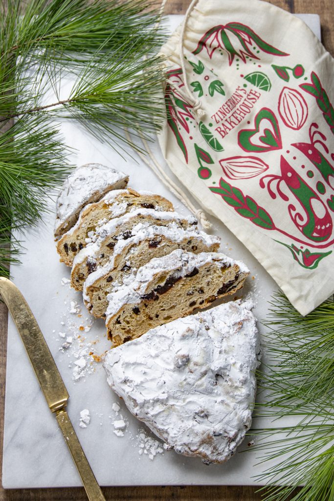 an overhead view of a loaf of stollen on a marble surface with some slices cut, a decorative muslin bag next to it, a gold knife, and some greenery