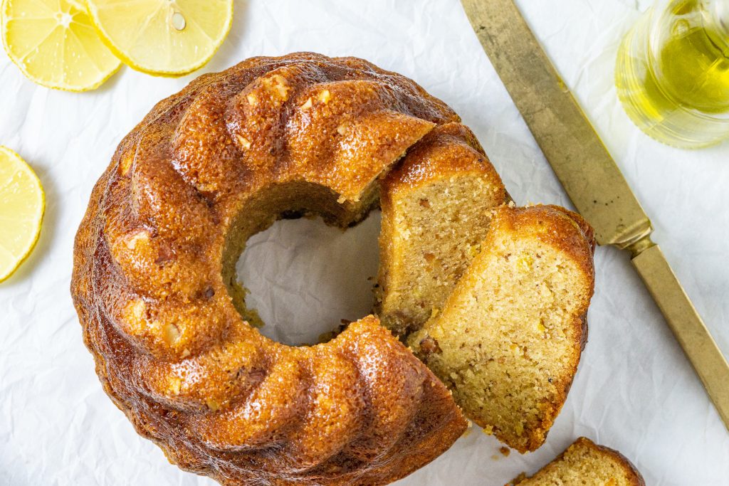 overhead view of an olive oil cake with a couple of slices cut, lemon slices, a gold knife and a glass container of olive oil