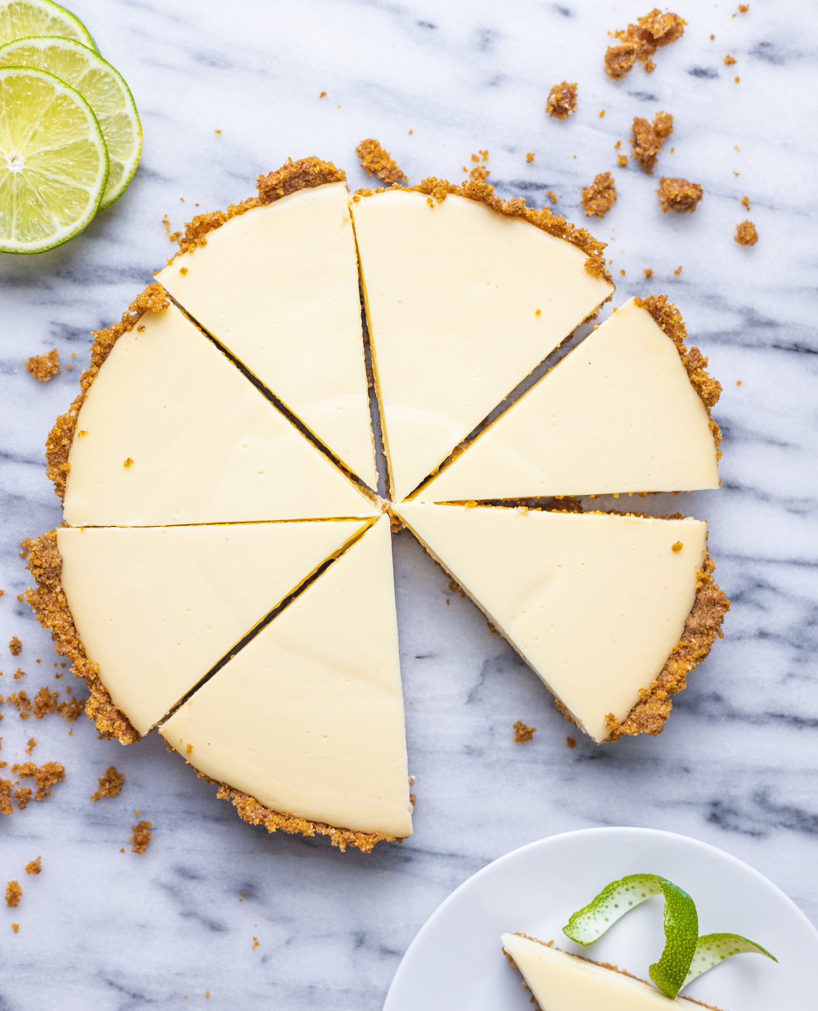 an overhead view of a Key Lime Pie on a marble surface, cut into 8 slices, with one slice on a white plate, mostly out of view