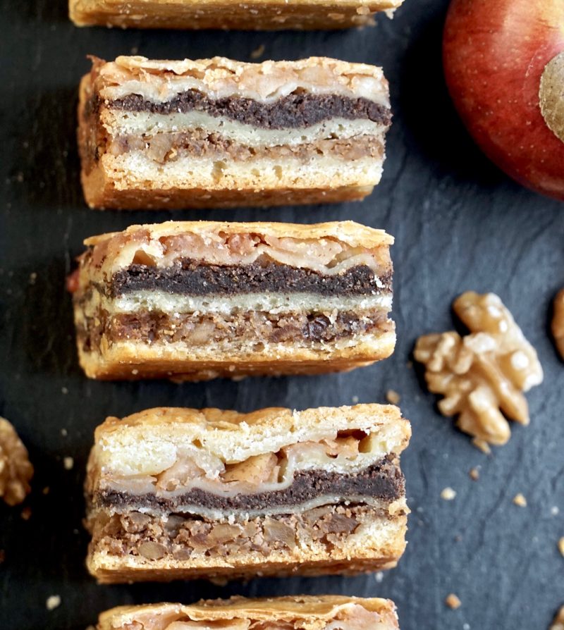 an overhead view of multiple squares of flodni, set on their sides, to show off the layered fillings, on a slate surface, with a partial view of an apple and walnuts