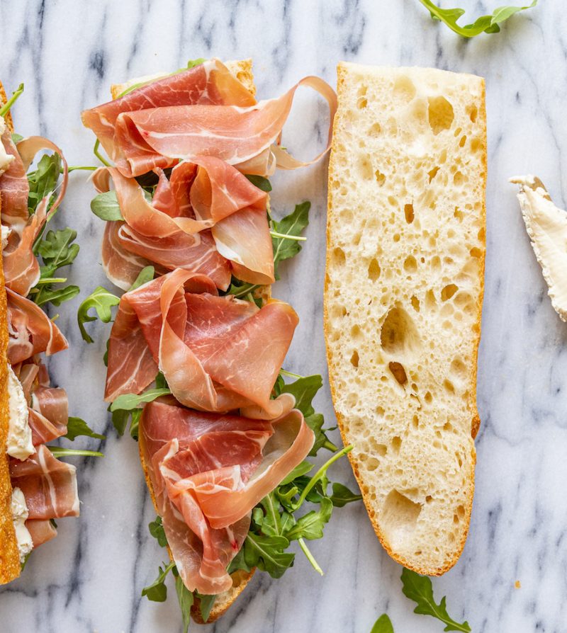A French baguette being used to make sandwiches with prosciutto, brie, and arugula