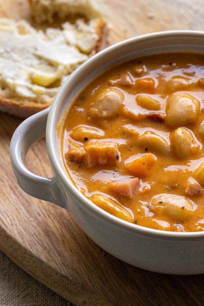 A bowl of Gabor's Butter Bean and Ham soup on a wooden surface with a slice of buttered bread visible behind it