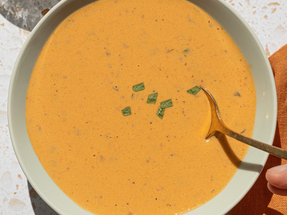 A close-up of John's Roasted Reds soup in a mint green bowl with a gold spoon and a hand holding it.