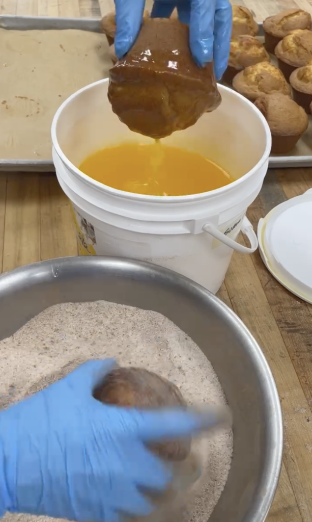 A muffin being dunked into a bucket full of melted butter in one gloved-hand, while another muffin is being rolled in a bowlful of cinnamon sugar by a 2nd gloved hand
