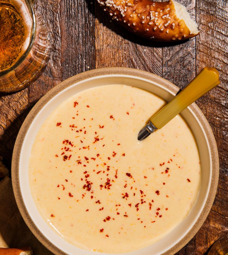 a bowl of cheddar ale soup on a wooden table with a glass of beer and a soft pretzel visible around it
