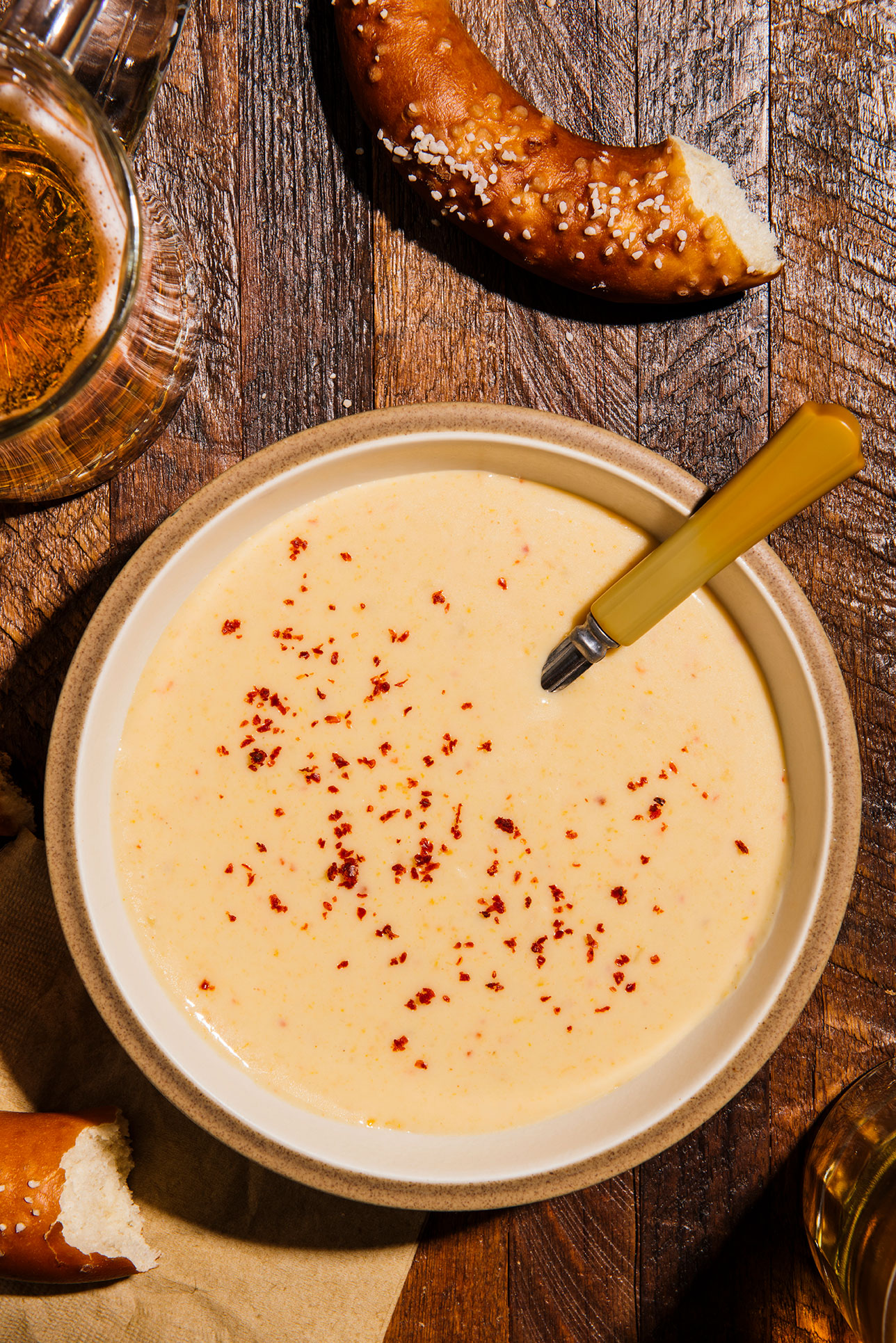 a bowl of cheddar ale soup on a wooden table with a glass of beer and a soft pretzel visible around it