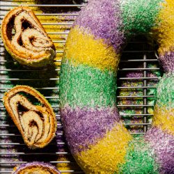 a new orleans king cake on a cooling rack with slices of king cake next to it
