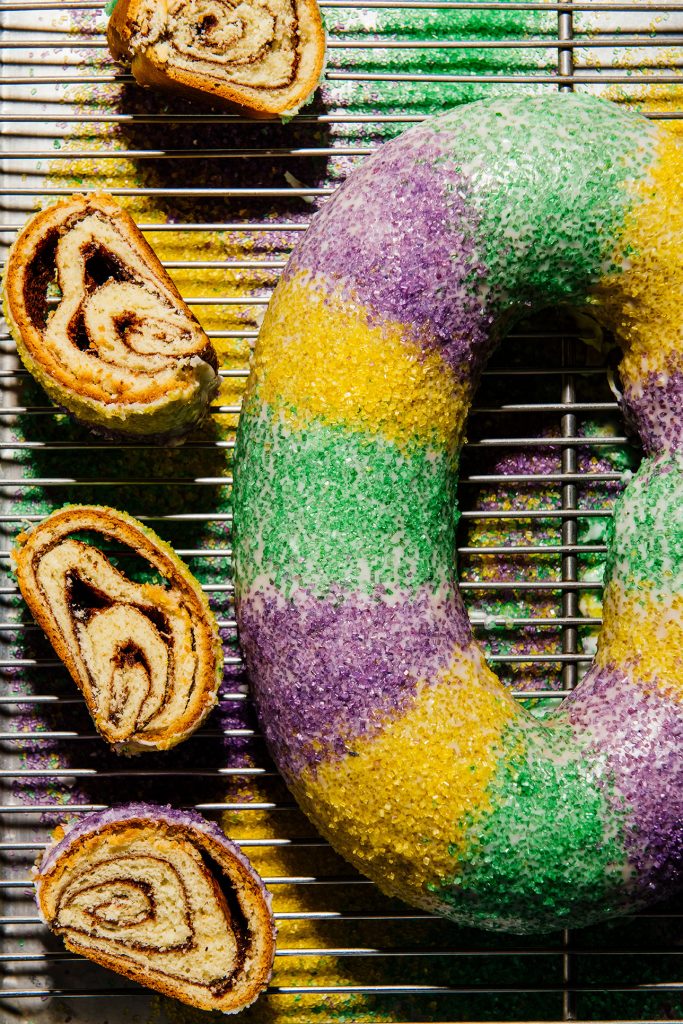 an overhead view of a New Orleans King Cake on a metal cooling rack with 3 slices of King Cake arranged to the left of it; green, purple, and yellow sanding sugar is visible on the tray beneath the cooling rack