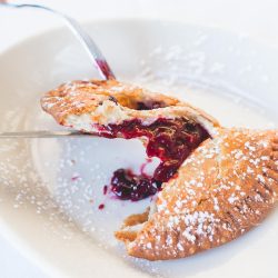 a fried hand pie filled with cherry on a white plate, cut in half. a fork and knife hold up one portion of it.