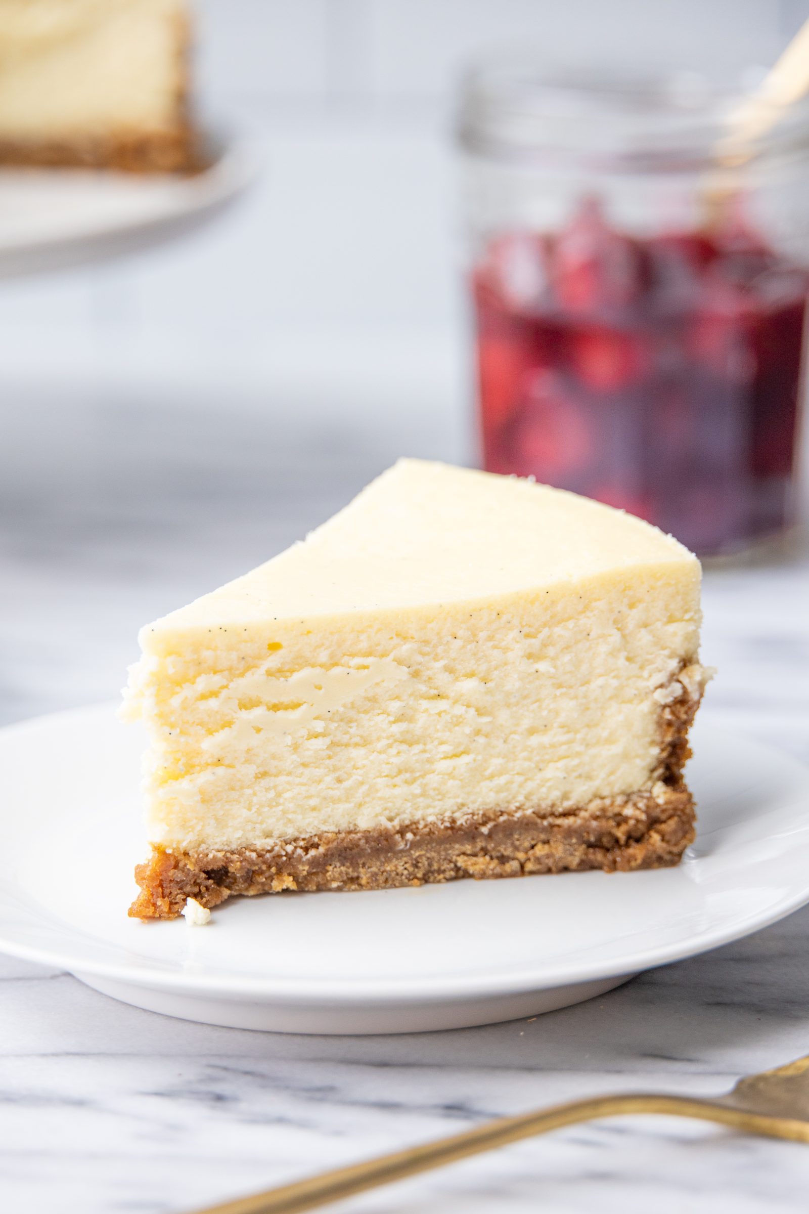 a slice of classic cheesecake on a white dessert plate, in front of a jar of cherries