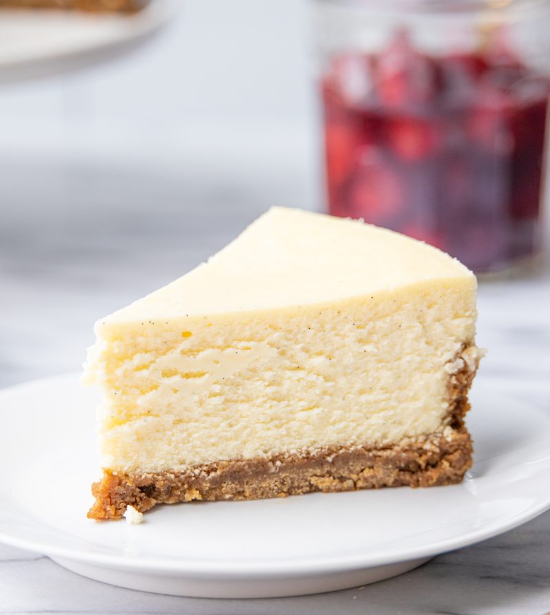 a slice of classic cheesecake on a white dessert plate, in front of a jar of cherries